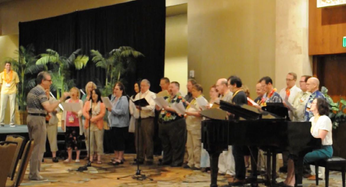 The choir singing beside a grand piano in front of palms and a black curtain in Hawaii.