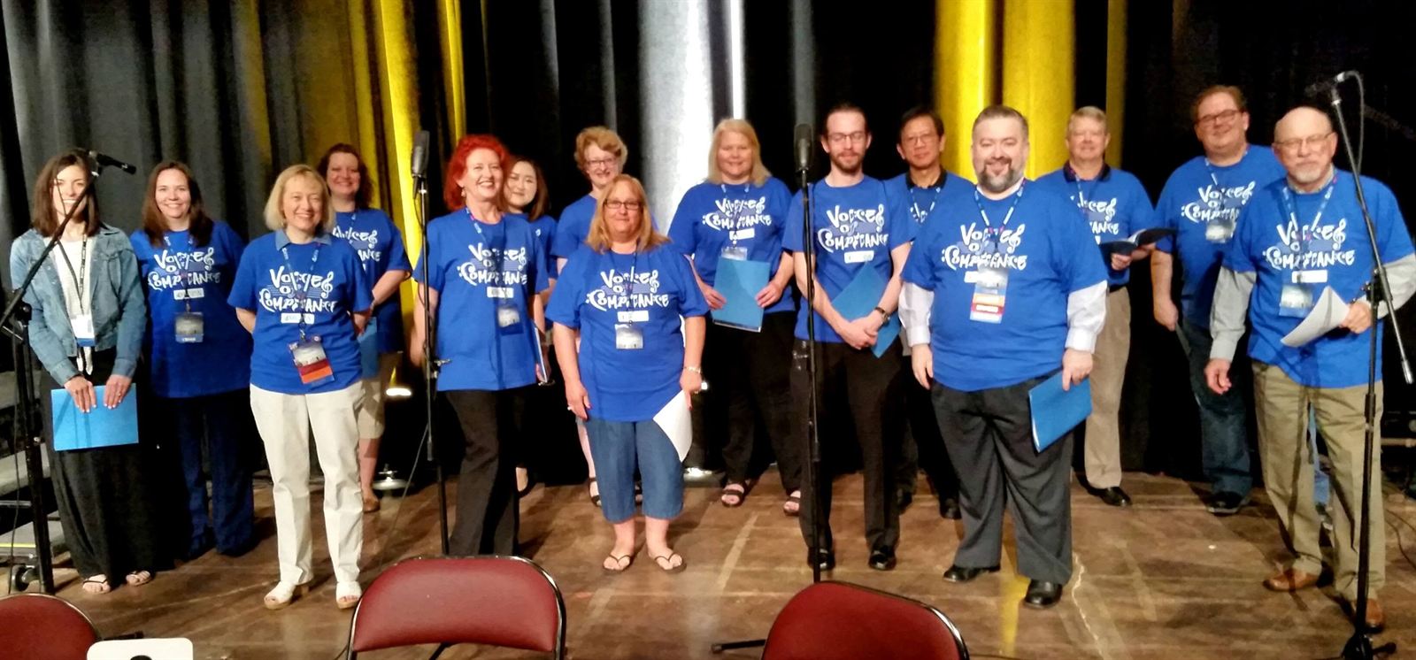 The choir lined up on stage in Voices of Compliance shirts, holding blue folders.