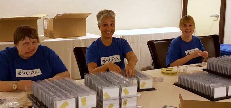 Three ICPA volunteers in blue ICPA shirts behind the conference registration desk, sorting rows of badge holders